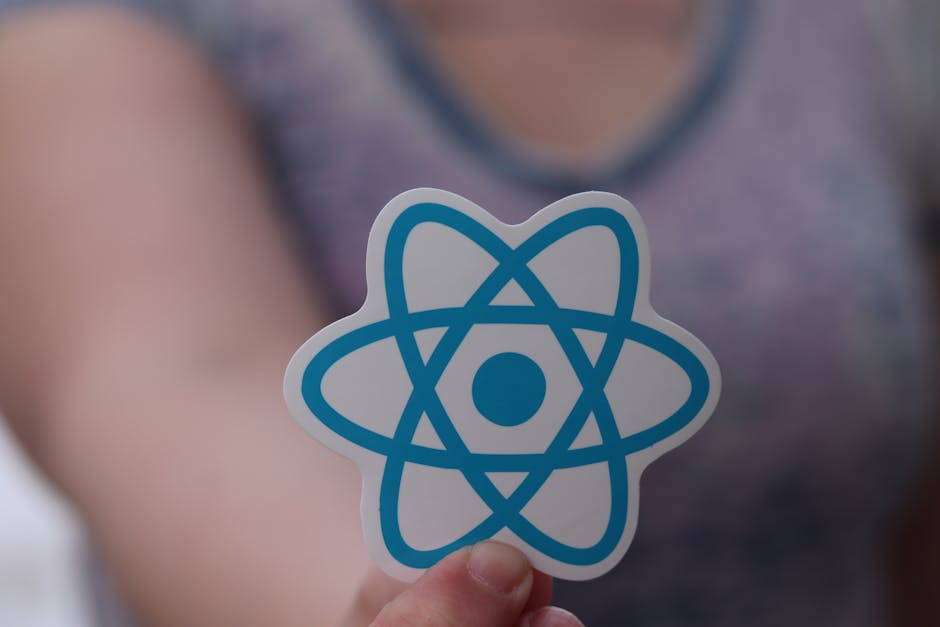 A person holding a sticker with a blue atomic symbol design in front of their torso, which features a grey sleeveless top. The background is blurred, focusing on the sticker, and the lighting appears