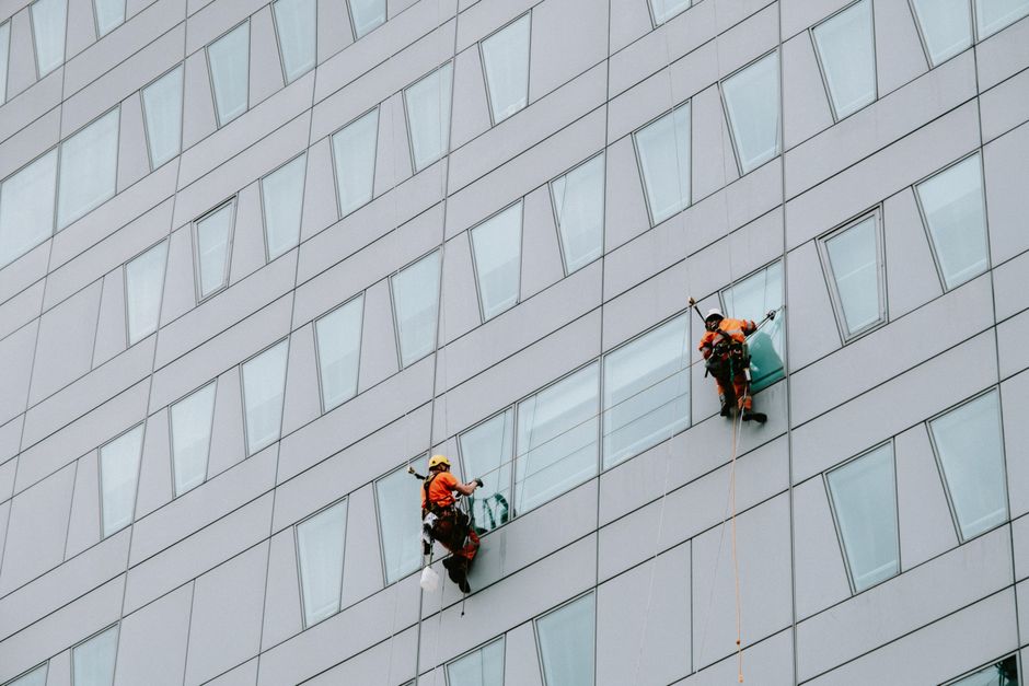 Two professional window cleaners from Archway Carpet Cleaners are suspended on harnesses and ropes, cleaning the large glass panes of a commercial building with a spray and squeegee. They wear protect
