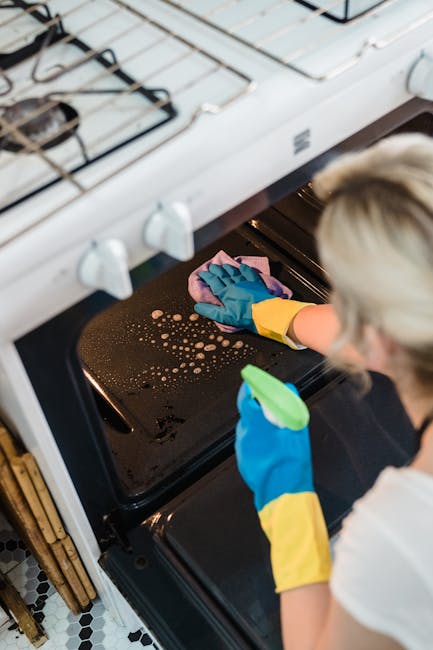 A person wearing blue rubber gloves and a white shirt is cleaning the black stovetop of an oven with a pink cleaning cloth, focusing on removing food splatters. The oven's interior is visible with a m