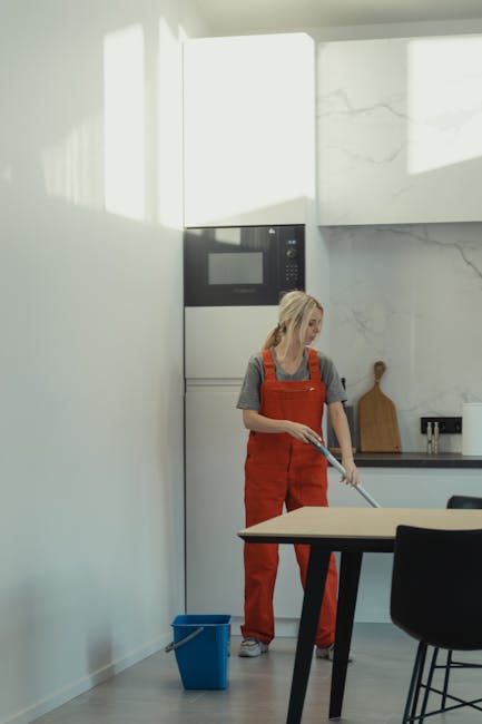 A person wearing blue rubber gloves and a white shirt is cleaning the black stovetop of an oven with a pink cleaning cloth, focusing on removing food splatters. The oven's interior is visible with a m