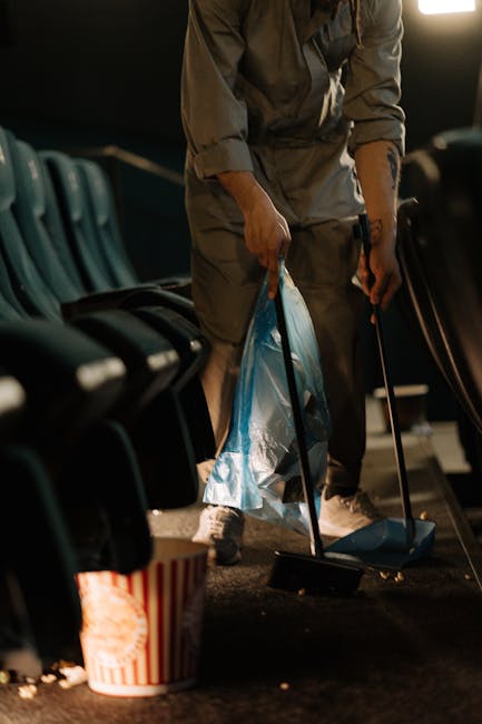 A person performing surface cleaning in an indoor space, using a broom and dustpan to sweep up debris from the carpeted floor, which is surrounded by a row of dark green upholstered seats. The area is